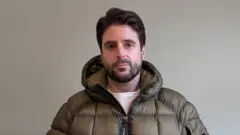 Alex Grossomanides has brown hair, brown eyes and a beard. He is wearing an olive green puffer coat and looking at the camera while standing in front of a white wall indoors.