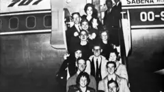 يقف أشخاص خارج طائرة يحملون لافتة مكتوباً عليها "رابطة التزلج الفني الأمريكية" في صورة بالأبيض والأسود