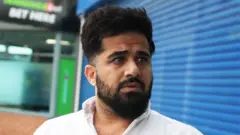 Close-up of Twana Jamal who has dark hair and a full beard, wearing a white collared shirt, standing outdoors in front of a blue metal shutter. He is turned slightly to one side, not facing directly toward the camera. In the background, there is a building entrance with glass doors and a partially visible green sign.