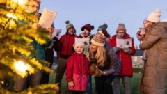 A group of people of different ages, standing beside a decorated Christmas tree, wrapped in jackets and hats, with their eyes staring on leaflets at the back of which a Christmas tree picture can be seen