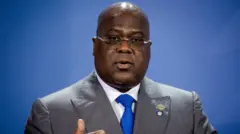 Perezida wa DR Congo Félix Tshisekedi yafotowe ubwo yari ari mu kiganiro n'abanyamakuru i Berlin mu Budage, ku itariki ya 27 Kanama (8) mu 2021, mu nama yahuje itsinda ry'ibihugu bikize cyane rya G20 n'Afurika. Yambaye indorerwamo, ikositimu iri hagati y'amabara ya khaki n'ikigina, ishati y'umweru na karuvati y'ubururu, akoze ikimenyetso n'ikiganza. 