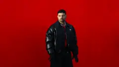 Węgierski raper Ádám Schwartz, znany jako Beton.Hofi, pozuje na tle czerwonego tła. Ma na sobie ciemną kurtkę nałożoną na czerwoną koszulkę piłkarską oraz czarne rękawiczki i patrzy w dal.