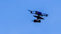 A fibre-optic drone flies in a clear blue sky across the border from Lebanon into Israel. It has a spool of fibre optic wire handing under a chassis, supported by four propeller blades around a blue rectangular battery.