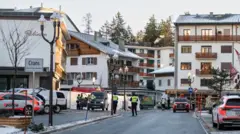 白い壁の建物が並び「Crans」という標識に書かれた道沿いに、警察車両が数台停車している。また、蛍光黄色のベストを着た警官が数人立っている