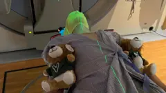 Anthony Perriam está deitado em uma cama de hospital com dois ursinhos de pelúcia ao seu lado. Ele está usando uma máscara porque vai realizar um exame para detectar câncer