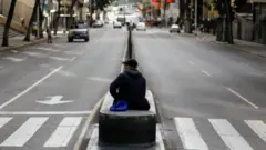 Каракас, 5 января 2026 года: на барьере, разделяющем проезжую часть улицы, прямо рядом с пешеходным переходом сидит мужчина в бейсболке. Машин на улице немного, людей тоже