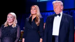 يظهر في الصورة بالترتيب من اليسار إلى اليمين (المتحدثة باسم البيت الأبيض، كارولين ليفيت، والسيدة الأولى، ميلانيا ترامب، والرئيس الأمريكي، دونالد ترامب) جميعهم يرتدون ملابس رسمية تليق بالاحتفال لونها أسود. ترامب يرتدي بدلة رسمية سوداء وربطة عنق كلاسيكية سوداء (باو تاي - بابيون). ليفيت "حامل" تضع يديها أسفل بطنها وتلبس فستاناً أسود اللون ولامع. وميلانيا ترتدي فستاناً أسود.