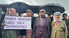 Dört kadın, geleneksel köy kıyafetleriyle yanyana duruyor, en soldakinin elinde Maden Yasasına Karşı Halk Dayanışması yazılı bir pankart var. Geri iki siyah şemsiye ve diğer köylüler flu. 