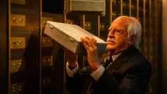 Ary Fontoura, de pele clara, com cabelos brancos penteados para trás e um bigode branco marcado. Ele veste um terno escuro, com camisa clara e gravata, e tem uma expressão concentrada enquanto segura uma caixa metálica em um banco.