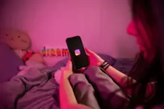 Jovem sentada na cama olhando para o celular. Na tela, aparece o aplicativo do Instagram
