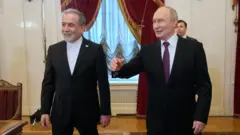 Abbas Aragachi (izq.) y Vladimir Putin fotografiados juntos en San Petersburgo. Ambos visten trajes oscuros y están de pie en un salón con cortinas ornamentadas