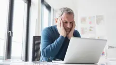 رجل في منتصف العمر يجلس متجهما أمام شاشة كمبيوتر "لابتوب"