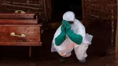 Umukozi wo kwa muganga yambaye impuzu zo kumurinda Ebola, iruhande rwe hari isandugu, mu buseruko bwa Repubulika ya Demokarasi ya Congo.