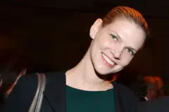 Nadia Marcinko está de frente para a câmera com a cabeça levemente inclinada, vestindo um blazer escuro sobre uma blusa verde e pequenos brincos.