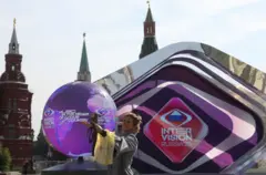 Uma mulher tira uma selfie na Praça Vermelha, em Moscou. Atrás dela, há um grande balão decorativo e um telão com as palavras INTERVISION RUSSIA'25 e o slogan “Música no coração do seu país”.