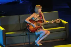 Taylor Swift sentada em um sofá no palco, tocando um violão de 12 cordas e cantando ao microfone durante uma apresentação ao vivo. Vestindo um figurino brilhante com franjas prateadas e sapatos de salto claros, ela aparece sob iluminação azul e amarela.