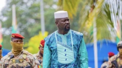 Le président Mamady Doumbouya en boubou bleu et vert avec un chapeau blanc, derrière lui se trouvent des militaires en béret rouge.