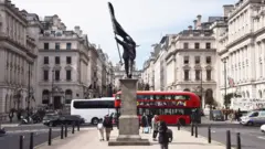 A grey statue of a man wearing a suit, walking forward off a plinth, carrying a flag that covers his face. A red London bus can be seen in th bcakground