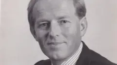 Retrato em preto e branco de John Smyth, usando camisa listrada com colarinho e terno escuro. Ele sorri para a câmera e aparenta ser amigável