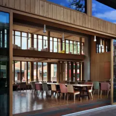 Uma grande porta de vidro aberta revela uma sala comum espaçosa e iluminada, com dois andares, revestida de madeira, com mesas e cadeiras de cores diferentes.