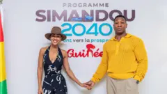 Meagan Good (à droite) vêtue d'une combinaison noir et blanc et d'un fedora marron, et Jonathan Majors (à gauche) vêtu d'un pull jaune et d'un pantalon beige se tiennent la main devant une pancarte sur laquelle on peut lire « Programme Simandou 2040 ». Un drapeau guinéen est déployé à l'extrême droite du cadre