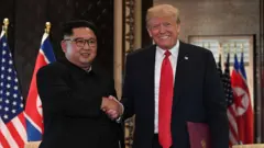 À esquerda, Kim Jong-un, em um terno escuro estilo “Mao”; à direita, Trump, de terno preto e gravata vermelha. Os dois líderes sorriem e apertam as mãos diante das bandeiras dos EUA e da Coreia do Norte