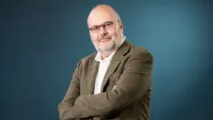 Eeconomista Branko Milanovic fotografado da cintura para cima, com os braços cruzados e ligeiramente virado de perfil. Ele veste um paletó marrom, camisa branca e usa óculos redondos, enquanto olha para a câmera com um leve sorriso