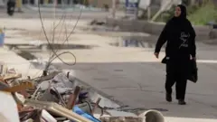 سيدة بلباس أسود وحجاب تحمل حقيبة سوداء وتسير قرب ركام مبنى متضرر من القصف