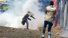 Umugabo uri mu myigaragambyo umaze gutera ikintu, inyuma ye hari umwotsi, imbere ye hari undi mugabo usa nk'urimo kumufotora