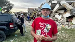 Ronnie Navarro con una franela roja y un casco azul y la pila de escombros atrás.