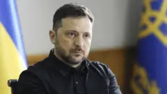 Volodymyr Zelensky sentado em uma cadeira, vestindo uma camisa preta e olhando para a direita da câmera. Ele está sentado diante de uma bandeira da Ucrânia.