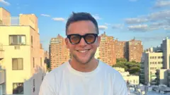 Retrato colorido de Andrew Greene, um homem branco jovem, usando uma camiseta branca, óculos de lente amarela e com uma cidade ao fundo