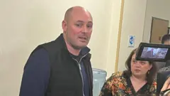 Ліворуч - у спортивній темно-синій кофті Руслан Магомедрасулов.На ньому також чорна жилетка. Праворуч - його адвокатка Олена Щербан у чорні кофті з квітчастим принтом. Вона звернена до нього. 