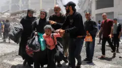 Família com pertences em meio a construções destruídas após bombardeio na Faixa de Gaza