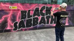 Um fã com uma camiseta do Black Sabbath tirando uma foto de um grande mural colorido do Black Sabbath em uma parede em Birmingham