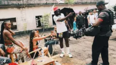 Meji Alabi, con un sombrero rojo, una camiseta blanca, pantalones cortos negros y calcetines y zapatillas blancas, sostiene una cámara y señala durante una sesión fotográfica. A su lado hay mujeres sentadas en tumbonas junto a un árbol delgado. Un camarógrafo, con una gorra negra y vestido con ropa oscura, está de pie al otro lado y el personal de producción, algunos con sombrillas negras, está detrás.