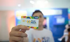 Jovem desfocado ao fundo segurando um cartão do programa Pé de Meia, que aparece em foco e no primeiro plano