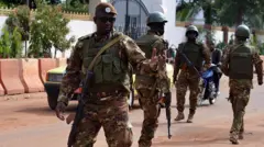 Four soldiers in frame, one dey wear glasses wit gun for im neck, one dey back di camera. Anoda bike man dey for di back