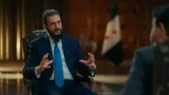 Şara oturmuş konuşurken elleriyle jest yapıyor. Muhabirin arkası dönük geride Suriye bayrağı da flu. 