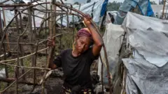 Umugore wambaye umupira w'umukara, ukenyeye igitenge, ufashe ku biti arimo kugonda akazi k'ibiti mu nkambi, inyuma ye hari izindi ngonde ziriho amahema zimeze nk'iyo ashaka kubaka