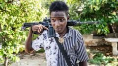 Um jovem membro de gangue de 14 anos com um fuzil AK-47 nos ombros e um medalhão dourado no pescoço patrulha as ruas do bairro Mariani, de Porto Príncipe (Haiti), em 6 de outubro de 2025, perto da Route Nationale 2, parcialmente tomada pelas gangues