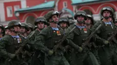 Um grupo de soldados armados, vestidos com uniformes verdes, marcha no desfile do Dia da Vitória, que marca o fim da Segunda Guerra Mundial, em Moscou, em 9/5/2026. Todos avançam com a perna esquerda. O soldado no centro olha para a frente e sorri amplamente; os demais olham de lado, em direção à câmera, com expressão mais séria