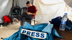 Мужчина сидит на стуле в палатке, глядя в телефон. На столе - бронежилет с надписью PRESS и бутылка воды 