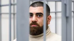 Маркарян в клетке в зале суде. У него густые брови, коротко стриженные волосы на голове, густые, но аккуратные усы и борода. Он одет в светлую водолазку