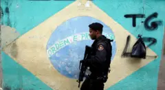 Policial passa na frente de bandeira do Brasil pintada em muro durante operação policial no Rio