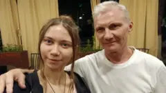 Седой мужчина в белой футболке держит руку на плече девушки, оба смотрят в камеру, слегка улыбаясь