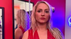 चेहऱ्यावर गंभीर भाव असलेल्या आणि कॅमेऱ्याकडे पाहणाऱ्या मेलचा फोटो. तिचे केस लांब, सोनेरी आणि नागमोडी आहेत. तिने व्ही-नेक लाल ड्रेस घातला आहे. ती आरशासमोर उभी आहे. छताला साखळी लटकलेली आहे आणि निऑन लाइट्स देखील फोटोमध्ये दिसत आहेत.