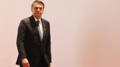 Bolsonaro aparece de terno e em pé na frente de fundo branco