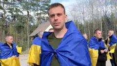 На відкритій місцевості стоїть група людей, багато з них загорнуті в синьо-жовті українські прапори. На передньому плані — чоловік з короткою стрижкою, також обгорнутий прапором, стоїть обличчям до камери.