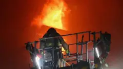 আগুন নেভানোর কাজ করছেন মইয়ের ওপর দাঁড়ানো একজন কমকল কর্মী